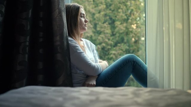 Sad pretty teenage girl sitting on windowsill. RAW video record.