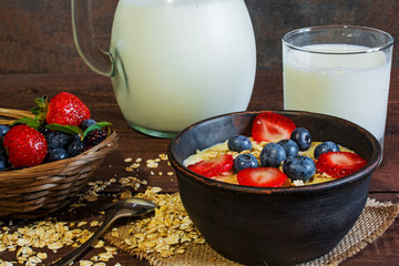 Milk in a jug and oatmeal porridge with glass of milk in a pottery bowl with fresh ripe berries