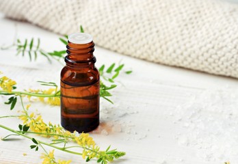 Bottle of essential oil, green herbs, sea salt scattered, beige cotton towel. Herbal extract for skincare. 