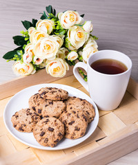 tray with chocolate chip cookies, cup of tea and flowers on wood