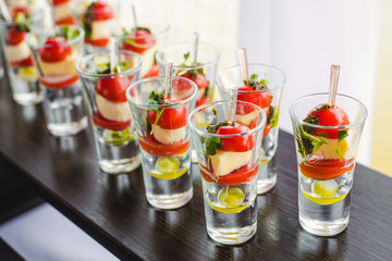 Catering for party. Close up of appetizers with cherry tomatoes, green olives, olive oil, cheese and spices in short glasses on wood brown table.