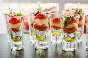 Schilderijen op glas Catering for party. Close up of appetizers with cherry tomatoes, green olives, olive oil, cheese and spices in short glasses on wood brown table. © Andrii Oleksiienko