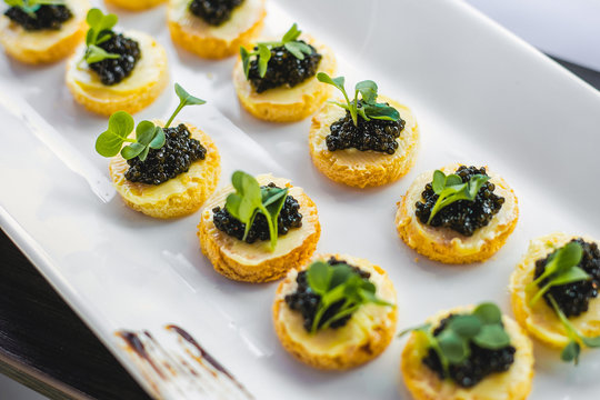 Close up of two raws of black fish caviar toasts on white plate on festive table. Catering for party or wedding celebration. Close up horizontal photo of expensive appetizers.