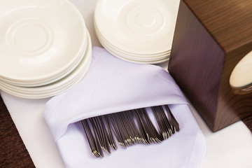 Catering equipment on table. Many forks or knifes in white clean napkin waiting for guests. White dessert plates on brown wood table. 