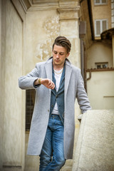 Elegant attractive young man outdoor wearing wool coat, in European city, looking at wrist watch