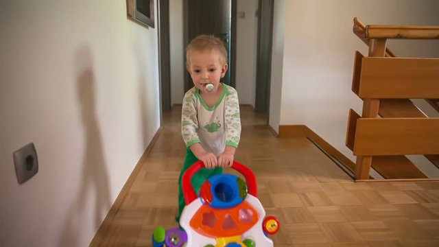 A Small Boy Is Pushing A Toy Trolley Across The Corridor And He Is Smiling. He's Having Cute Green Tights On And A Pacifier In His Mouth.
