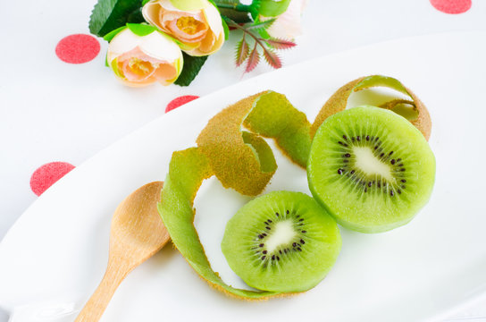 Peeled off kiwi fruit on white plate with skin and wooden spoon over red and white dots table cloth with small roses for decorating