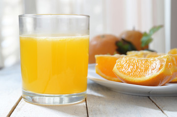 A glass of natural oranges served next to some slices on a white wooden table.