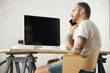 Close view on tattooed man in blank white t-shirt speaking on mobile phone while sitting on his working space with his vintage bicycle parked near, close to white brick wall