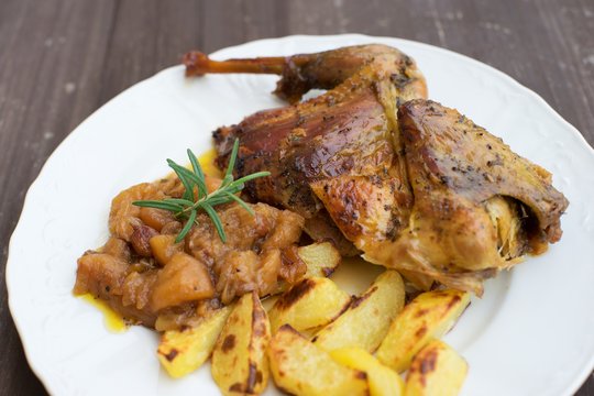 Roasted or baked Guinea fowl served with baked potatoes and sweet onion with apples and raisins. Meal spiced by rosemary and black pepper is served on the white plate and old rustic wooden table.