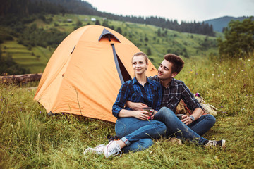 A couple of tourists in time of the hike, sit and relax outside the tent, admiring the beautiful mountain scenery. The guy hugs the girl. The concept of love, tenderness and recreation