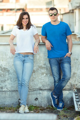 Young woman and man are standing on the rooftop terrace. They are leaning on the wall behind them, looking at camera and smiling.
