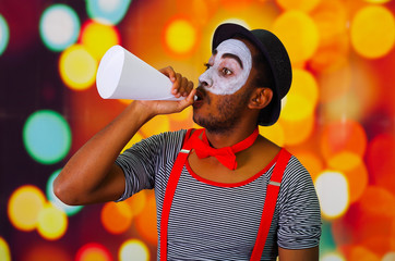 Pantomime man with facial paint posing for camera holding blowing horn, blurry lights background