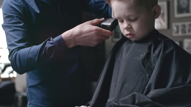 Barber brushing with comb and shaving hair on head of little boy with trimmer while he watching something on digital tablet