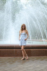 Attractive girl in short blue dress stands in front of fountain
