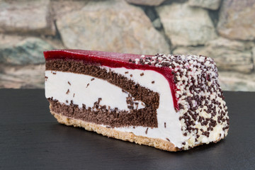 Ein Stück Kirsch-Sahne-Torte verziert mit Schokoladestreusel. Präsentiert auf einer Schieferplatte. Im Hintergrund eine Steinwand.