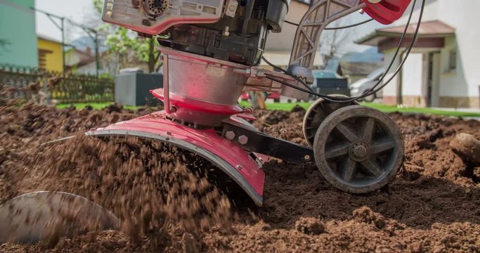 A senior gardener is helping out the family and he is plowing the soil with a cultivator in their garden. Close-up shot.
