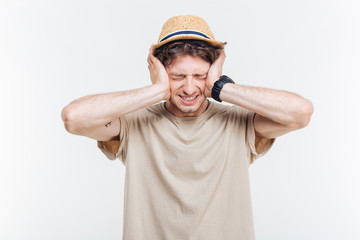 Frowning young man covered ears with hands over white background