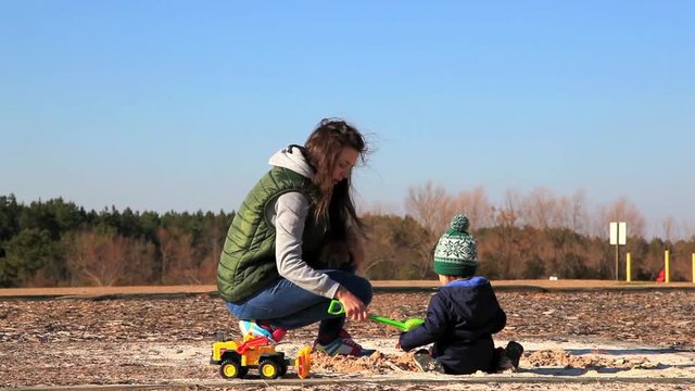 Mother and child toddler boy playing on the sand pit playground in the park with toys in cold sunny windy weather, woman helps small boy digging hole in sand