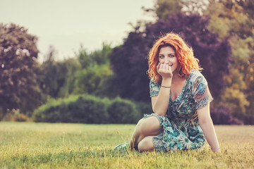 Girl with red curly hair and blue floral dress sitting on grass, covering her mouth with hand. Plenty of copy space