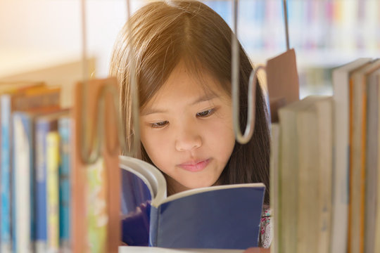 Little Asian Girl Reading A Book In Library At School