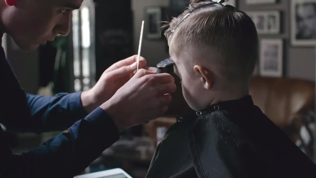 Barber cutting hair of little boy with trimmer while he using digital tablet