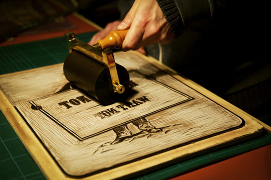 A signmaker rolling ink across the cut raised surface of linoleum preparing to print.