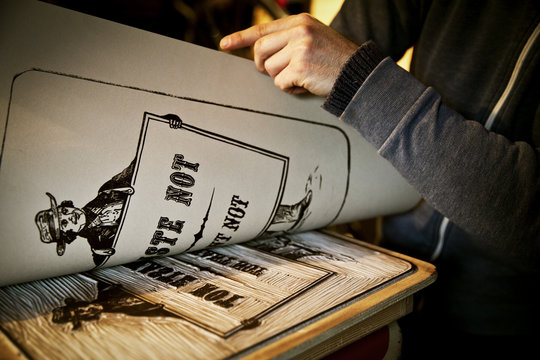 A signwriter lifting a sheet of imprinted card off a cutout linoleum surface, a woodcut. 