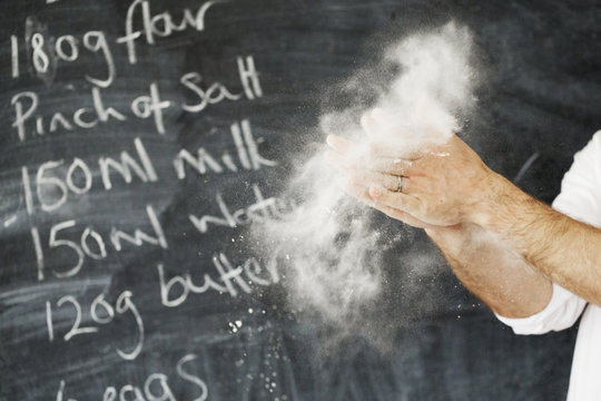 Close up of a baker standing in front of a blackboard, dusting his hands with flour.