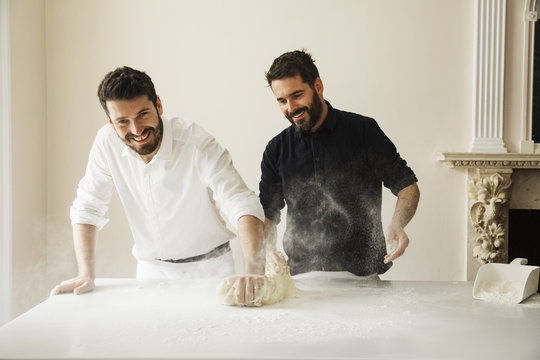 Two bakers standing at a table, kneading bread dough, dusting it with flour.