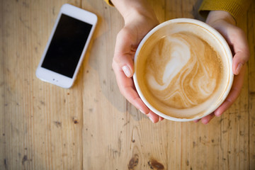 Above view of female hands holding hot cup of coffee  and with smart phone with on wooden table. Coffee break at work. Cappuccino while working.