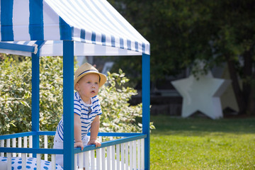 Obraz premium Adorable little toddler boy standing in the blue wood play, bed house and looking out of the window. Child playing outdoors on a sunny day.