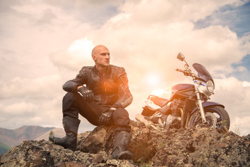Bald rider sits on a mountain next to  motorcycle and looks asi