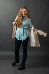 Stylish pretty blonde in blue blouse and dark blue pants dresses a beige coat on a dark gray background