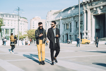 Obraz premium Two young handsome caucasian bearded brown hair businessmen walking in the city, both looking downward and tapping the screen of smartphone handhold and chatting- business, technology, work concept