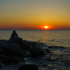 Man sitting on the stones at the seashore and watching beautiful sunset