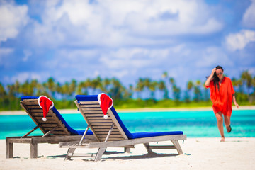 Two loungers with red Santa Hats on tropical white beach with turquoise water. Young woman walking on the beach
