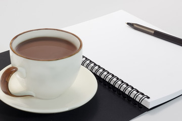 Blank notebook on white desk background with a pen on top and a coffee cup on the left.