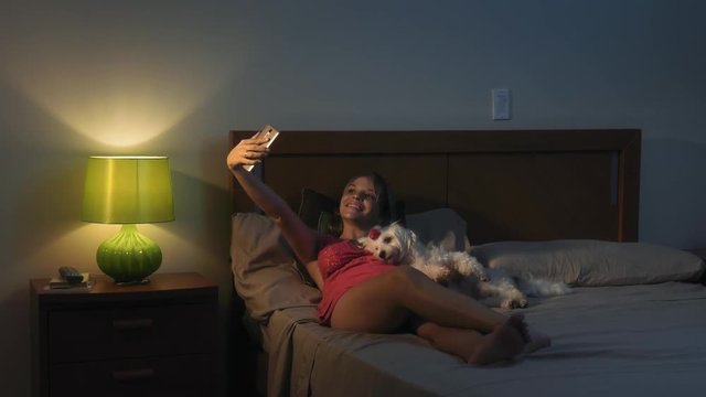 People and pets. Young woman taking selfie portrait with mobile phone at night. She is lying on bed, relaxing and embracing her small dog.