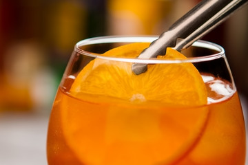 Tongs holding slice of orange. Glass filled with cocktail. Traditional recipe of aperol spritz. Sweet wine and fruit.
