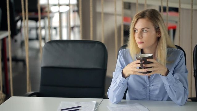 Pretty blonde woman sits at table in office and drinks coffee.