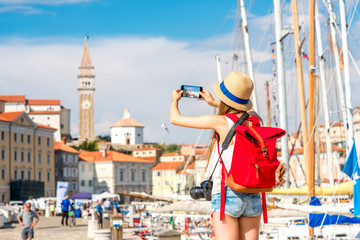 Young female traveler with hat and backpack photographing with phone piran town. Traveling in Slovenia