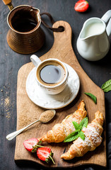 Breakfast set. Freshly baked croissants with strawberry, mint leaves and cup of coffee on wooden board served with pitcher and copper coffee pot over dark wooden backdrop, top view, selective focus