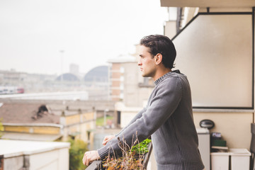 Half length of young handsome man standing on a balcony outdoor, overlooking, pensive - serious,...