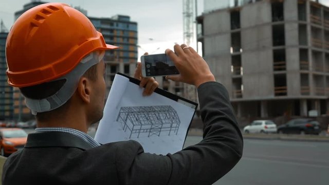 Slow motion. smart young attractive construction engineer with the smart phone taking pictures of objects on a construction site with beams and crane. Text tablet, orange helmet. Back close-up.