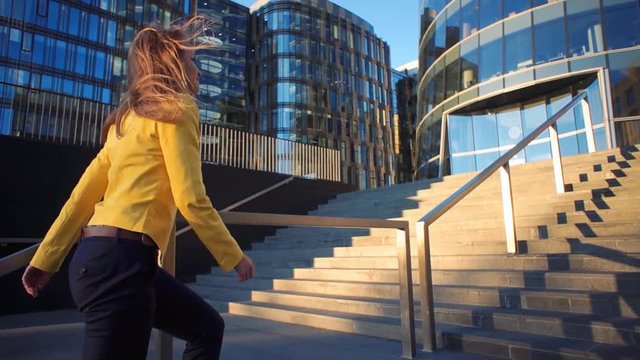 Young beautiful girl in a yellow jacket coming up the stairs. Business woman walking to work in the business center. Bright sunlight in the morning. Cinematic style video. Windly weather.