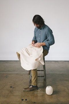 A man sitting on a stool, using string and hand making techniques to create an art piece.