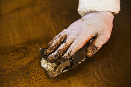 Close up of a carpenter wearing protective gloves, applying varnish onto a wooden surface with a cloth.