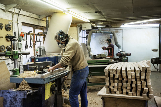 Man Standing In A Carpentry Workshop, Wearing A Respirator And Hearing Protector, Working On A Piece Of Wood.