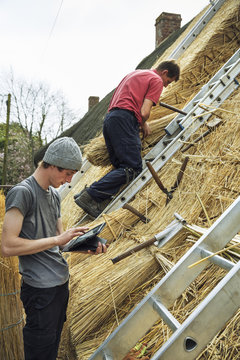 Two thatchers thatching a roof, one using a digital tablet the other standing on a ladder.